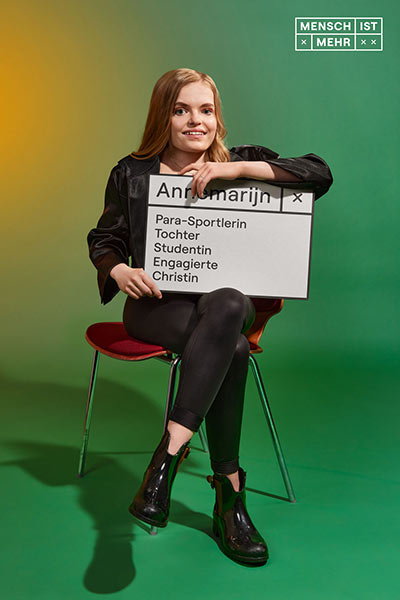 Eine junge Frau mit blondem Haar und schwarzen Klamotten sitzt auf einem Stuhl. Auf ihrem Schild steht: Annemarijn, Para-Sportlerin, Tochter, Studentin, Engagierte, Christin