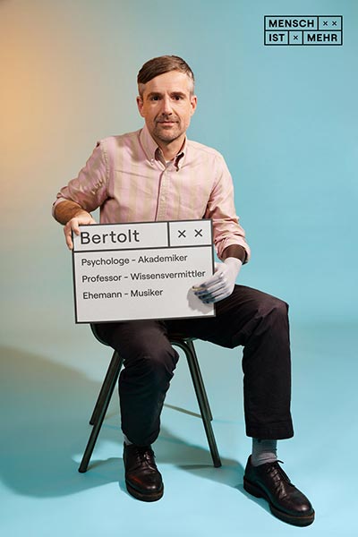 Ein Mann mit Hemd und einem Schild sitzt auf einem Stuhl. Er hat eine Handprothese. Auf dem Schild steht: Bertolt, Psychologe, Professor, Wissensvermittler, Ehemann, Musiker