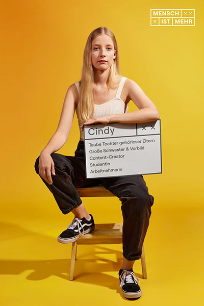 Eine junge Frau mit blondem Haar sitzt auf einem Hocker und schaut neutral. Auf ihrem Schild steht: Cindy, taube Tochter gehörloser Eltern, große Schwester und Vorbild, Content-Creator, Studentin, Arbeitnehmerin