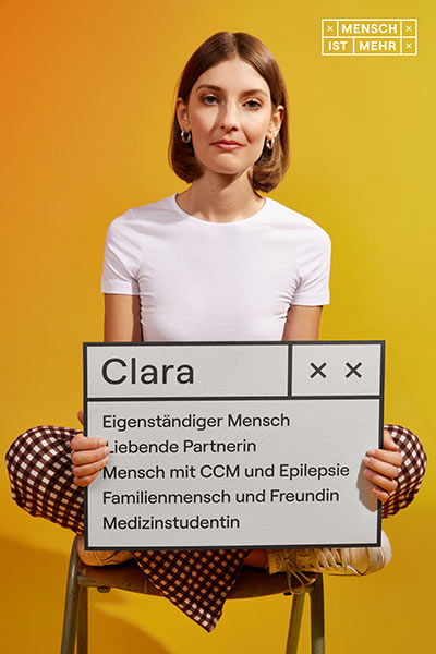 Eine elegante Frau mit schulterlangem Haar sitzt im Schneidersitz. Auf ihrem Schild steht: Clara, Eigenständiger Mensch, liebende Partnerin, Mensch mit CCM und Epilepsie, Familienmensch und Freundin, Medizinstudentin
