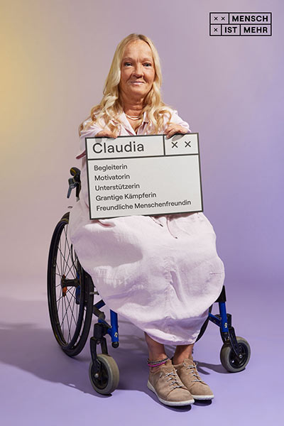 Eine Frau mit blondem Haar sitzt schmunzelnd in einem Rollstuhl. Mit kurzen Armen hält sie ein Schild: Claudia, Begleiterin, Motivatorin, Unterstützerin, Grantige Kämpferin, freundliche Menschenfreundin