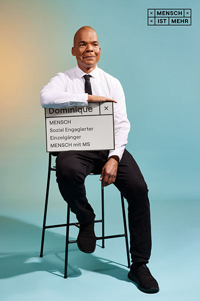Ein Mann mit Hemd und Krawatte schmunzelt sitzend in die Kamera. Auf seinem Schild steht: Dominique, Mensch, Sozial Engagierter, Einzelgänger, Mensch mit MS
