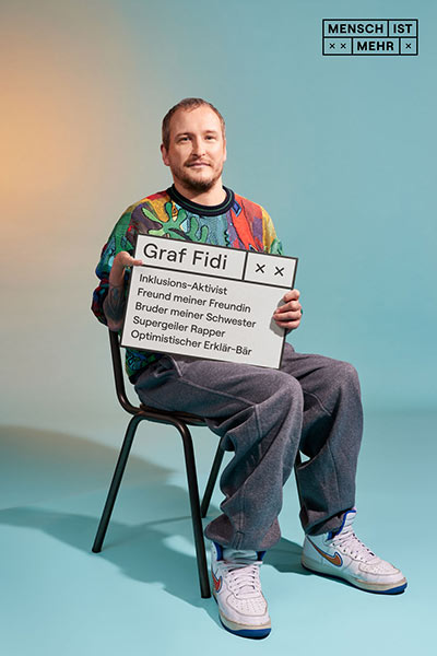 Ein Mann mit buntem Pullover sitzt auf einem Stuhl und schaut freundlich in die Kamera. Er hat sechs Finger, auf seinem Schild steht: Graf Fidi, Inklusions-Aktivist, Freund meiner Freundin, Bruder meiner Schwester, Supergeiler Rapper, Optimistischer Erklär-Bär