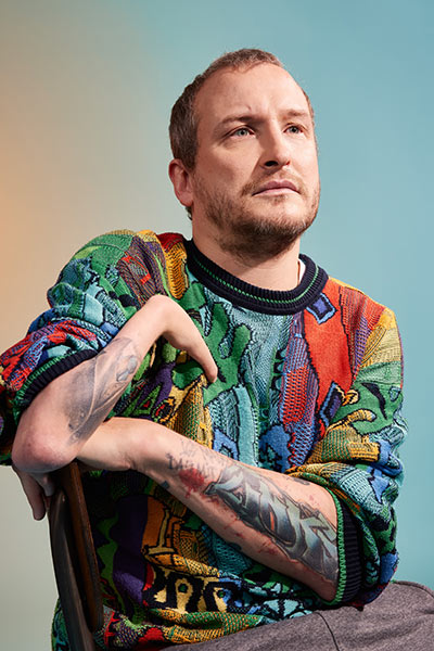 Ein Mann mit hochgekrempeltem Pullover und Tattoos auf den Armen schaut visionär in die Ferne. 