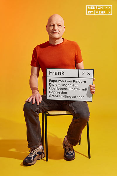 Ein Mann mit Glatze und orangenem Shirt sitzt auf einem Stuhl und schaut selbstbewusst in die Kamera. Auf seinem Schild steht: Papa von zwei Kindern, Diplom-Ingenieur, Überlebenskünstler mit Depression, Grenzen-Eingesteher