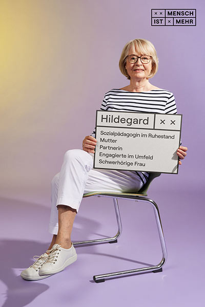 Eine junge Seniorin mit hellem Haar lächelt sitzend in die Kamera. Auf ihrem Schild steht: Hildegard, Sozialpädagogin im Ruhestand, Mutter, Partnerin, Engagierte im Umfeld, schwerhörige Frau