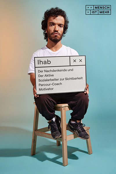 Ein junger Mann mit Locken sitzt auf einem Hocker und schaut verträumt. Er ist kleinwüchsig. Auf seinem Schild steht: Ihab: Der Nachdenkende und Der Aktive, Sozialarbeiter zur Sichtbarkeit, Parcour-Coach, Motivator