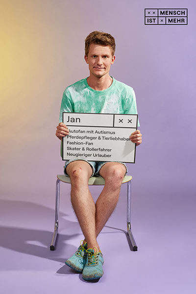 Ein junger Mann in Shorts schaut neutral. Auf seinem Schild steht: Jan, Autofan mit Autismus, Pferdepfleger und Tierliebhaber, Fashion-Fan, Skater und Rollerfahrer, Neugieriger Urlauber