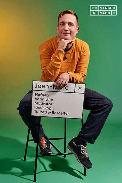 Ein junger Mann mit gelbem Pullover sitzt auf einem Hocker und schaut keck in die Kamera. Auf seinem Schild steht: Jen-Marc: Hofnarr, Vermittler, Motivator, Kindskopf, Tourette-Beseelter