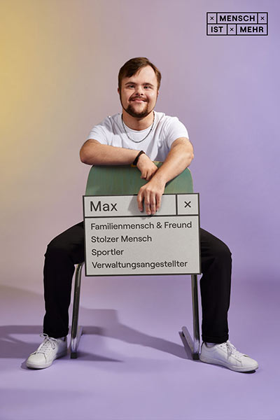 Ein junger Mann mit Bart sitzt lässig auf einem Stuhl. Auf seinem Schild steht: Max, Familienmensch, Freund, stolzer Mensch, Sportler, Verwaltungsangestellter