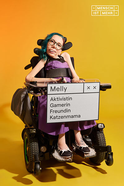 Eine Frau mit bunten Haaren und lila Kleid lächelt. An ihrem E-Rollstuhl hängt ein Schild: Melly, Aktivistin, Gamerin, Freundin, Katzenmama