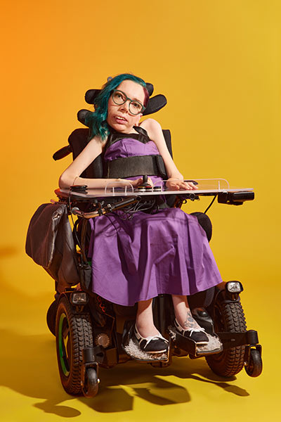 Eine Frau mit bunten Haaren und Brille schaut nach Vorne. Ihre Hände liegen auf einem Bedienpanel ihres E-Rollstuhls.