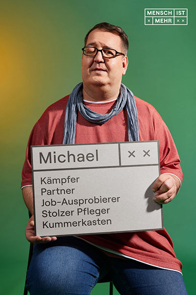 Ein Mann mit Schal und rotem Shirt schaut verschmitzt. Ein Auge ist zu. Auf seinem Schild steht: Michael, Kämpfer, Partner, Job-Ausprobierer, Stolzer Pfleger, Kummerkasten