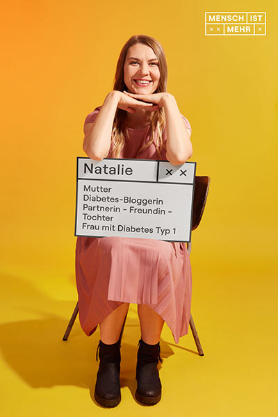 Eine Frau mit langem Haar strahlt und stützt den Kopf auf ihre Hände. Auf ihrem Schild steht: Natalie, Mutter, Diabetes-Bloggerin, Partnerin, Freundin, Tochter, Frau mit Diabetes Typ 1