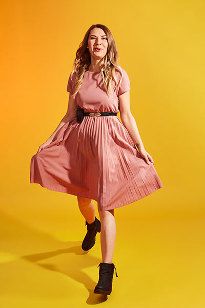 Eine Frau lacht und lässt ihr rosa Kleid flattern, während sie einen Schritt nach vorne geht.