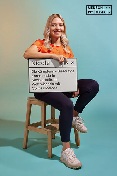 Eine junge Frau mit blondem Haar sitzt sportlich auf einem Hocker und lacht. Auf ihrem Schild steht: Nicole, Die Kämpferin, die Mutige, Ehrenamtlerin, Sozialarbeiterin, Weltreisende mit Colitis ulcerosa
