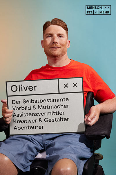 Ein Mann mit braunem Haar und rotem Shirt schaut selbstbewusst. Er sitzt in einem Rollstuhl. Auf seinem Schild steht: Oliver, Der Selbstbestimmte, Vorbild und Mutmacher, Assistenzvermittler, kreativer und Gestalter, Abenteurer