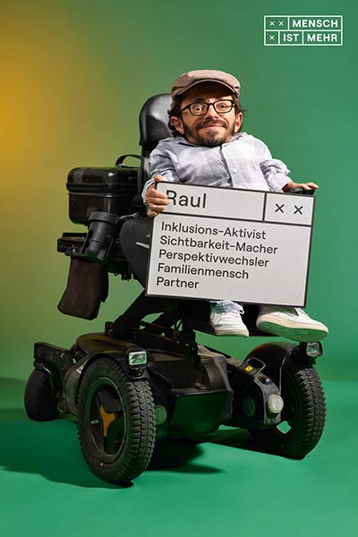 Ein Mann mit einer Schiebermütze und Brille schmunzelt in die Kamera. Auf seinem E-Rollstuhl sitzend hält er ein Schild: Raul, Inklusions-Aktivist, Sichtbarkeit-Macher, Perspektivwechsler, Familienmensch, Partner