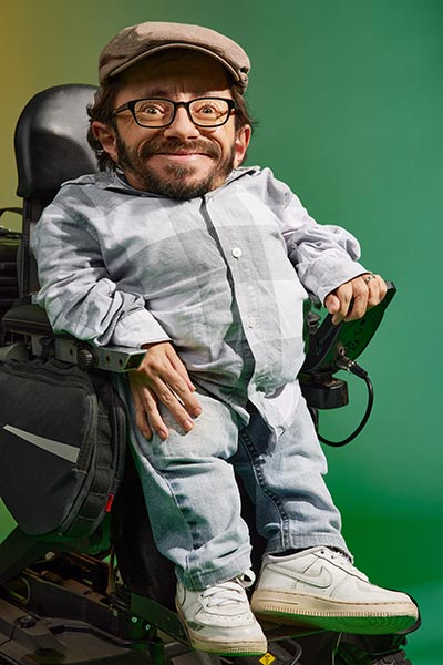 Ein Mann mit Schiebermütze, Bart und Brille grinst. Er trägt ein Hemd und Turnschuhe.