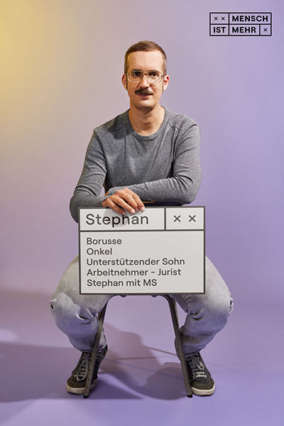Ein Mann mit Schnauzer und Brille schaut keck in die Kamera. Auf seinem Schild steht: Stephan, Borusse, Onkel, unterstützender Sohn, Arbeitnehmer, Jurist, Stephan mit MS