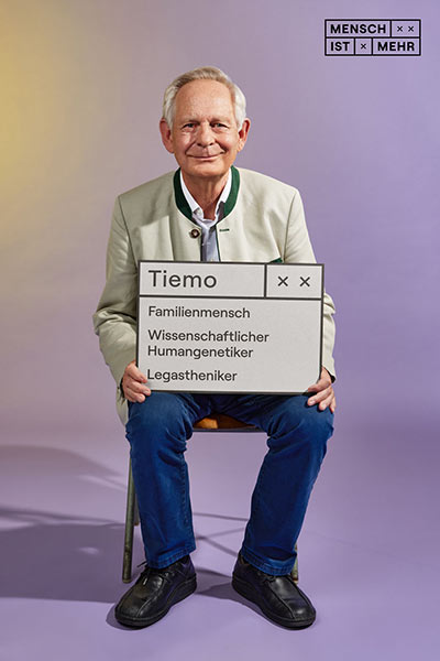 Ein älterer Herr mit bayrischer Jacke lächelt freundlich in die Kamera. Auf seinem Schild steht: Tiemo, Familienmensch, Wissenschaftlicher Humangenetiker, Legastheniker